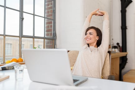 Happy relaxed young woman sitting in her kitchen with a laptop in front of her stretching her arms above her head and looking out of the window with a smile-1